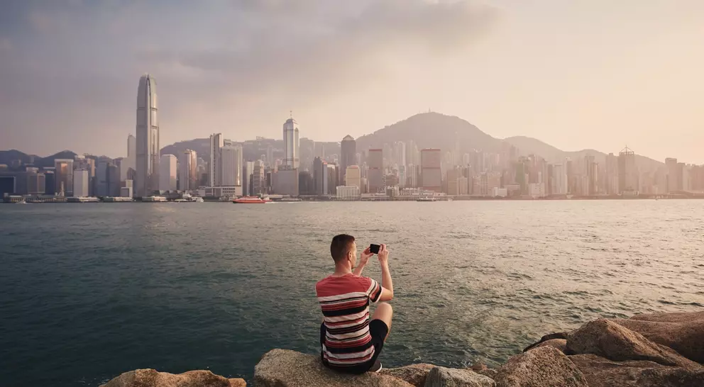 A person sitting on rocks by the water, taking a photo of the Hong Kong skyline at sunset.