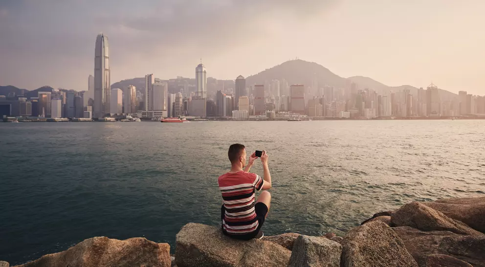 A person sitting on rocks by the water, taking a photo of the Hong Kong skyline at sunset.
