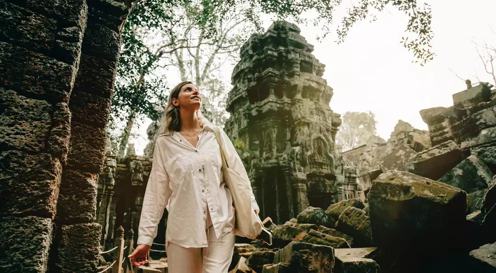 A woman in white clothing explores ancient ruins surrounded by overgrown trees and stone structures.