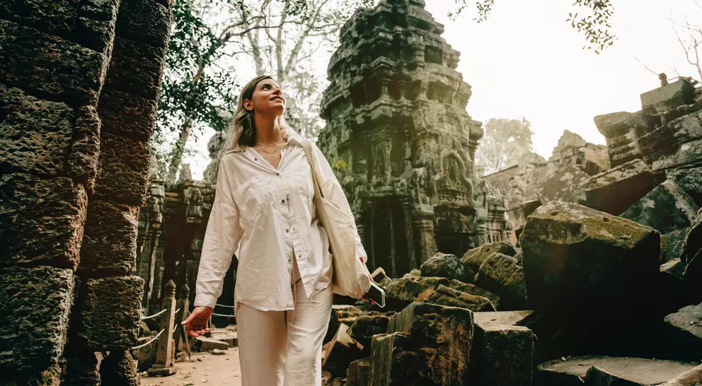 A woman in white clothing explores ancient ruins surrounded by overgrown trees and stone structures.