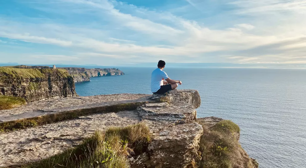 A person sits on a rocky cliff, overlooking a calm sea under a clear sky.