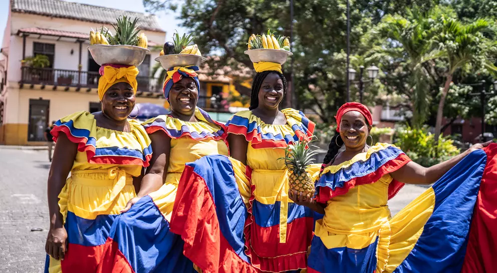 Four women in bright traditional dresses, each carrying a pineapple on their heads, pose happily outdoors.