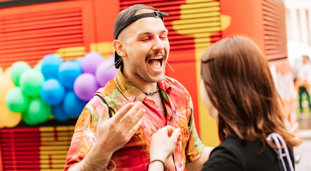 A person with colorful clothing and makeup joyfully interacts with another person against a vibrant, festive backdrop.