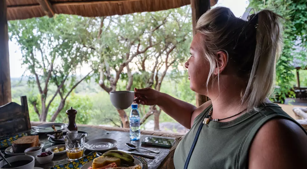 A person enjoying breakfast with fruits and drinks at an outdoor table, overlooking a scenic green landscape.