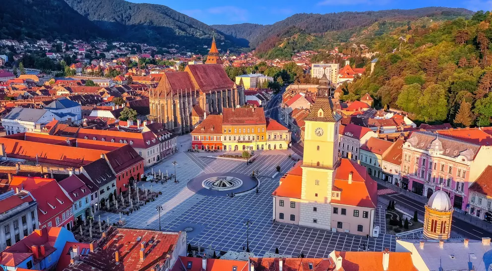 Aerial view of a historic town square with red rooftops, a clock tower, and mountains in the background.