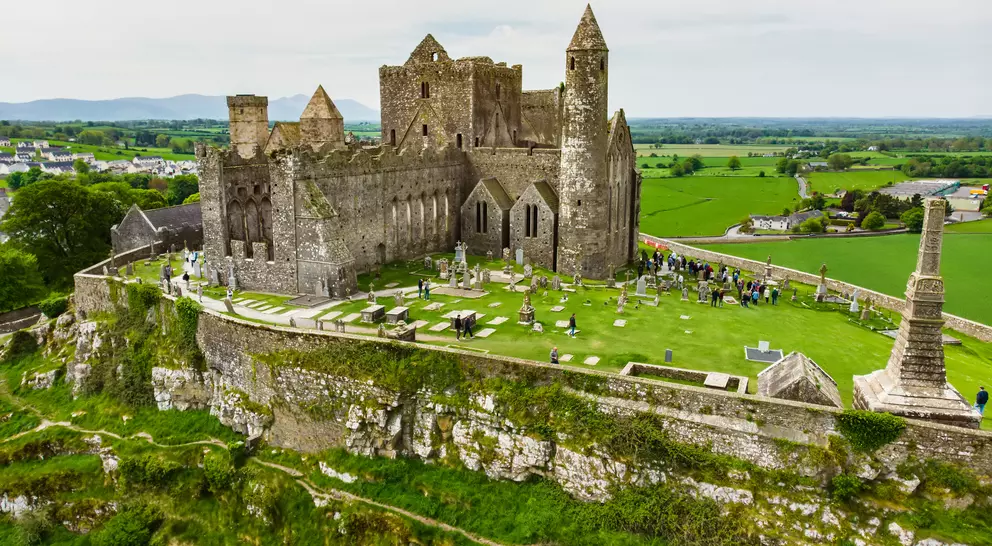Aerial view of a historic castle surrounded by green fields and gravestones on a hilltop.