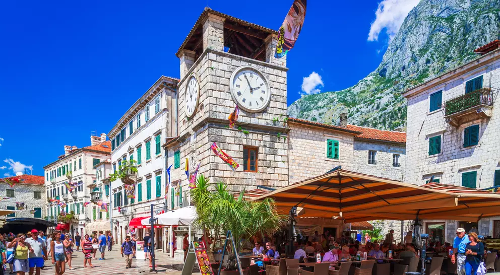 A bustling square in Kotor with a clock tower, cafes, and mountains under a clear blue sky.