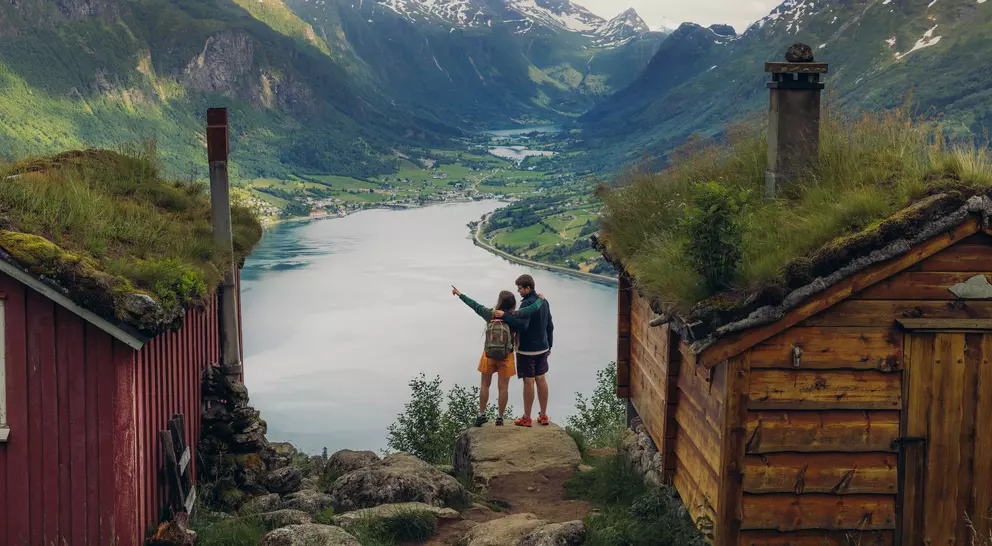 A couple stands between two wooden cabins, overlooking a scenic fjord surrounded by mountains.