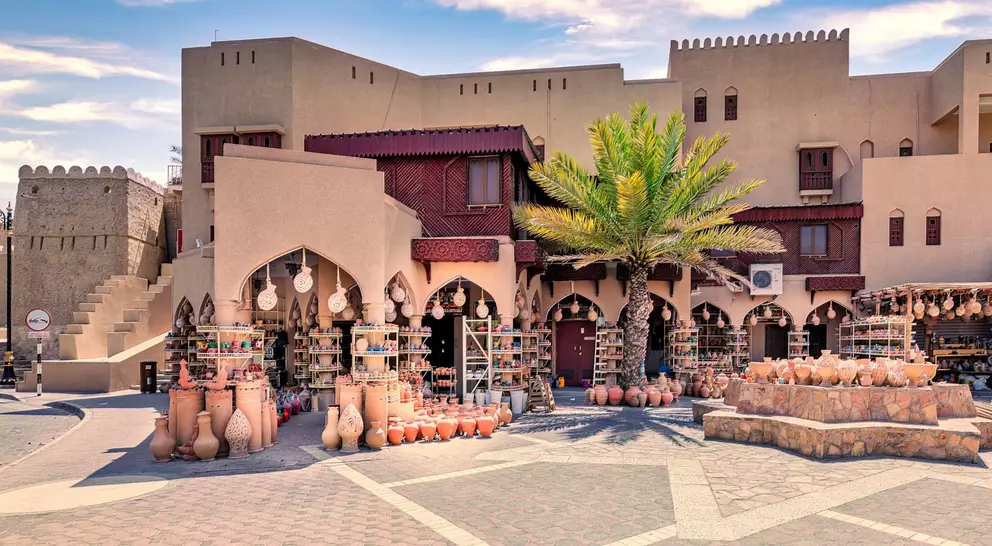 Market scene with clay pots for sale, palm trees, and traditional architecture under a blue sky.