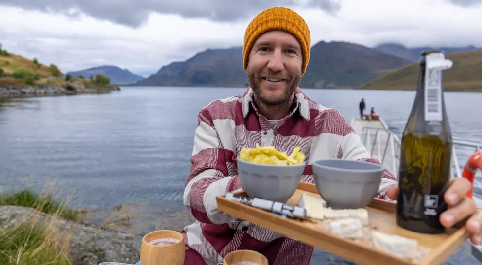 A smiling man in a striped shirt and orange beanie holds a serving tray with food and drinks by a calm lakeside.