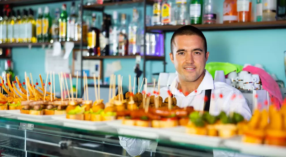 A smiling bartender stands behind a display of colorful tapas skewered with toothpicks, bottles of liquor visible in the background.