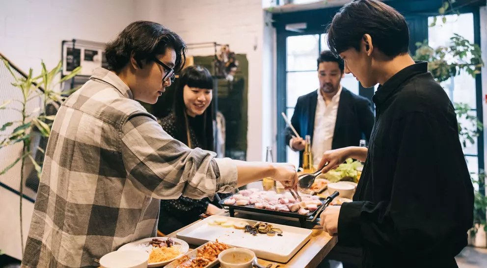 Group of friends enjoying a meal together, cooking at a table filled with various dishes and vibrant plants.