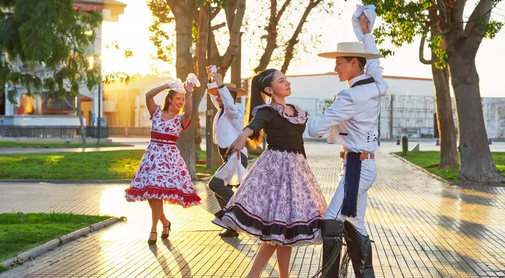 A couple dances in traditional attire outdoors, with more dancers in the background during sunset.