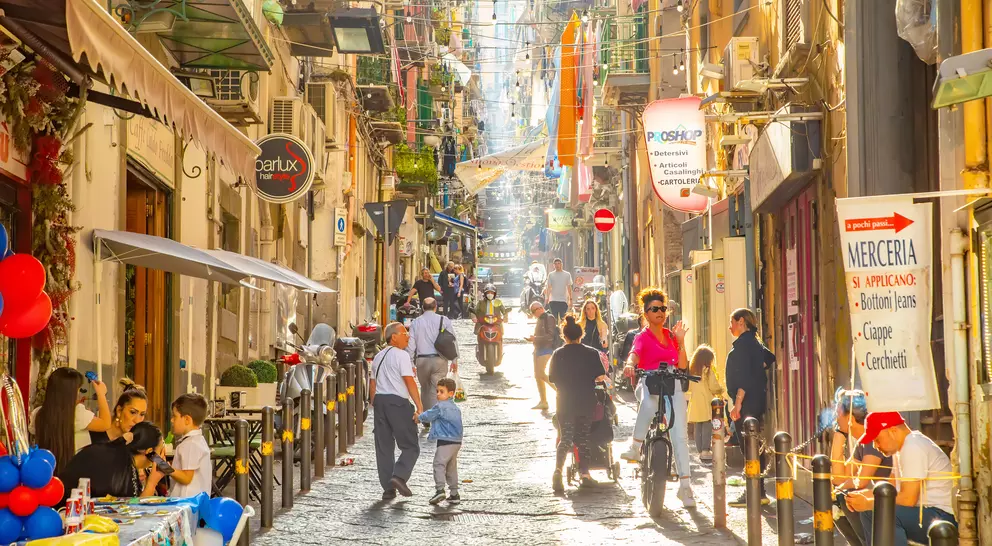 A bustling street scene with people walking, sitting at tables, and colorful decorations under bright sunshine.