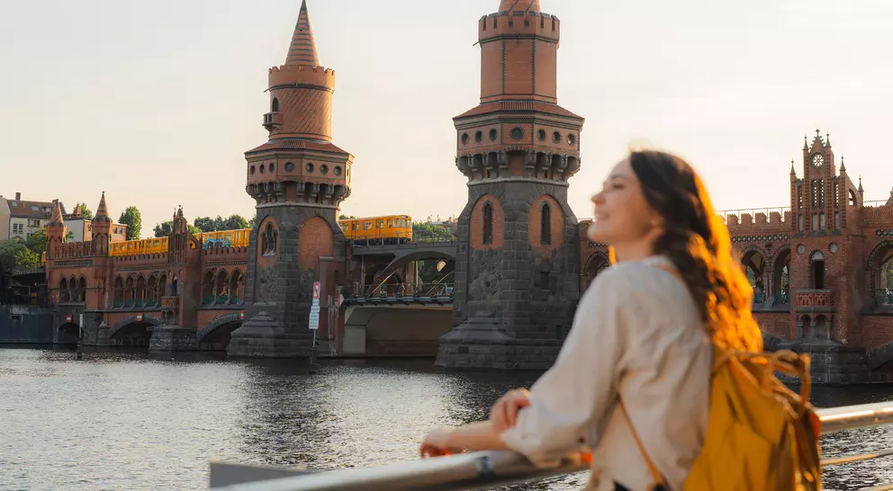 Young woman with a backpack gazes at a historic bridge with towers above a river during sunset.