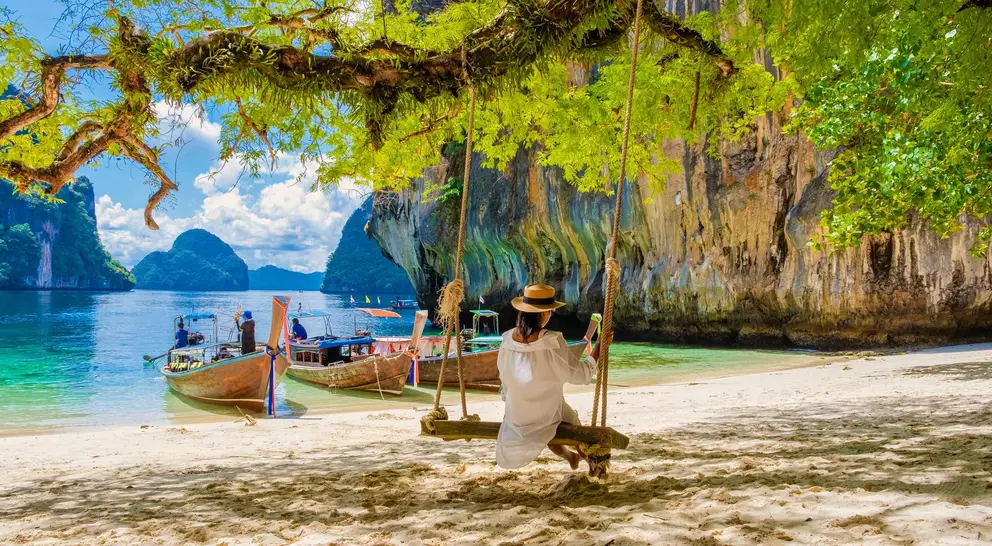 A person on a swing overlooking a tropical beach with boats and limestone cliffs in the background under a sunny sky.