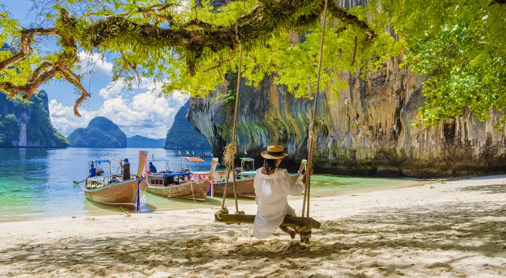 A person on a swing overlooking a tropical beach with boats and limestone cliffs in the background under a sunny sky.