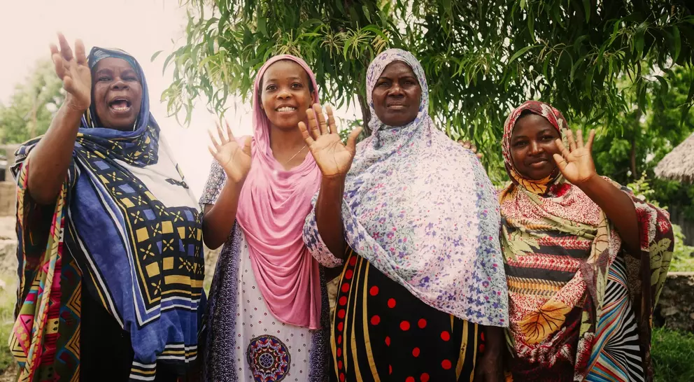 Four women in colorful dresses wave and smile, standing together under a tree in a vibrant outdoor setting.