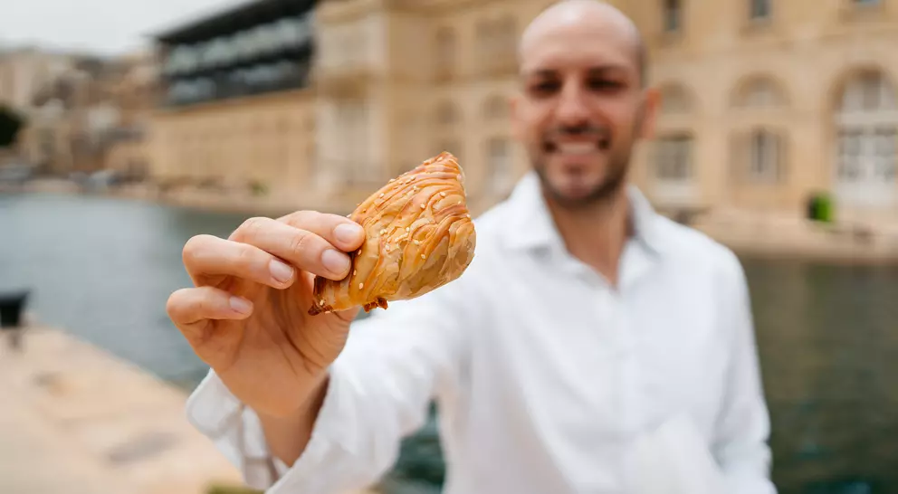 A man in a white shirt holds up a pastry by a calm waterway, smiling in front of a historic building.