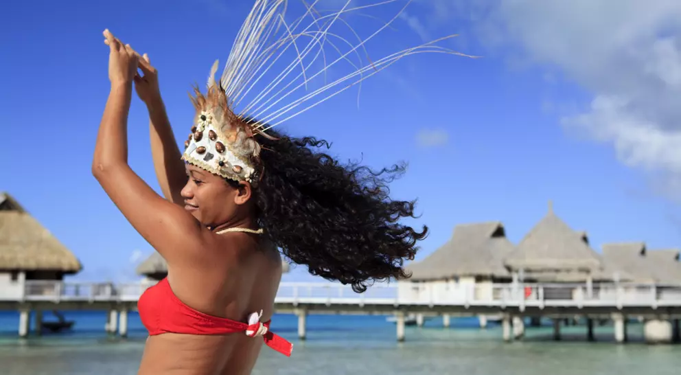 A dancer in a red bikini top and decorative crown sways against a tropical ocean backdrop with thatched huts.