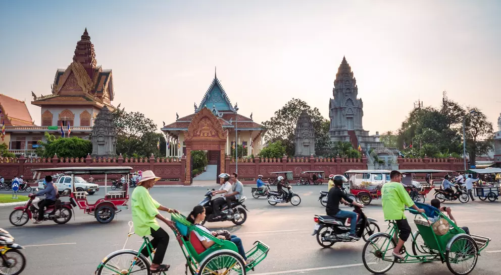 Traffic scene with rickshaws and motorbikes near a colorful temple complex at sunset.
