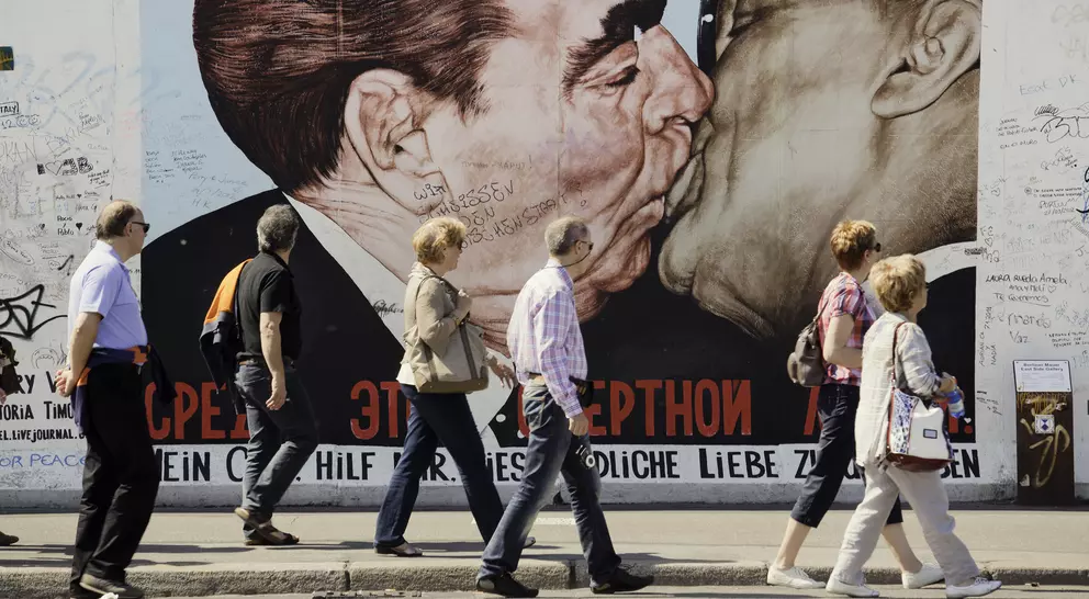 People walk by a large mural of two leaders kissing on a wall, with graffiti surrounding the artwork.