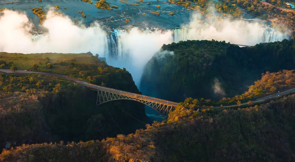 Aerial view of a waterfall with mist, featuring a bridge spanning a canyon surrounded by lush greenery.