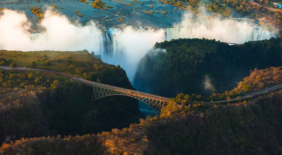Aerial view of a waterfall with mist, featuring a bridge spanning a canyon surrounded by lush greenery.