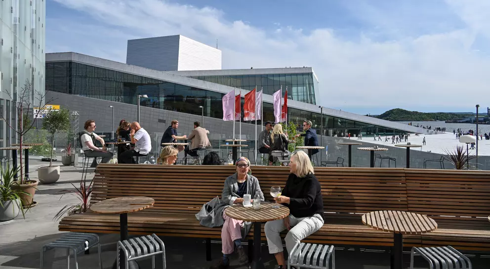 People relax on a sunny terrace outside a modern building, enjoying drinks and conversation. Flags flutter in the background.