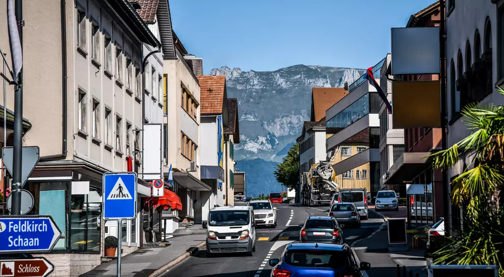 A street scene with shops, cars, and mountains in the background under a clear blue sky.
