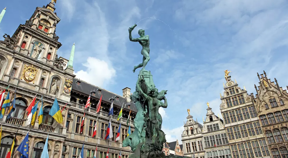 A bronze statue of a man atop a pedestal, surrounded by historic buildings and colorful flags under a blue sky.