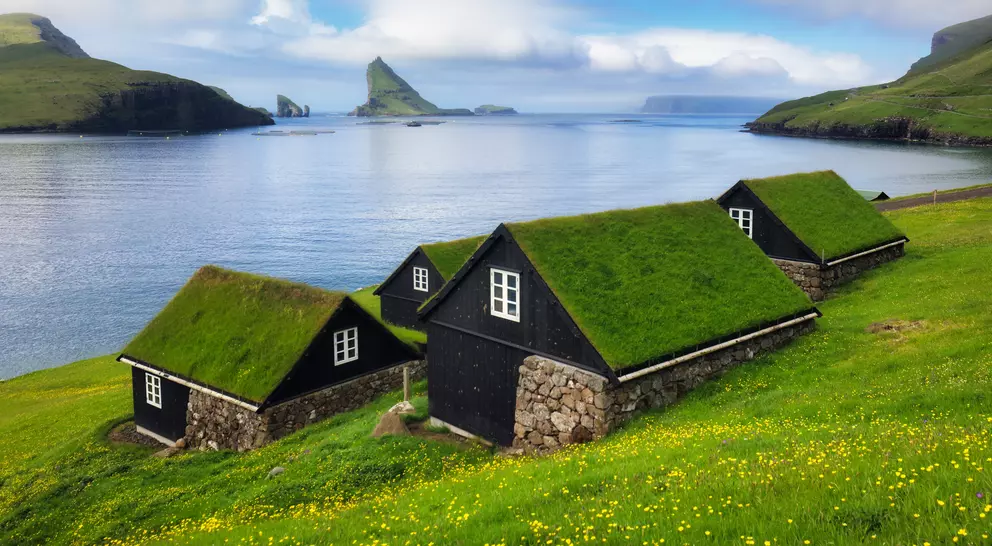 Black cottages with grass roofs on a green hillside overlooking a calm sea and distant rocky islands under a cloudy sky.