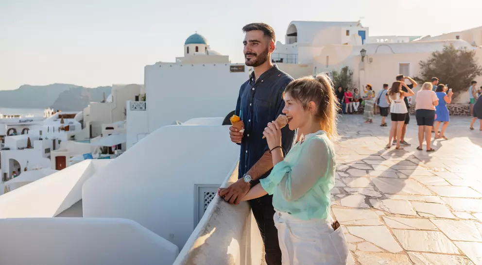 A man and a woman enjoy ice cream on a balcony, overlooking white buildings and people in a sunny coastal landscape.