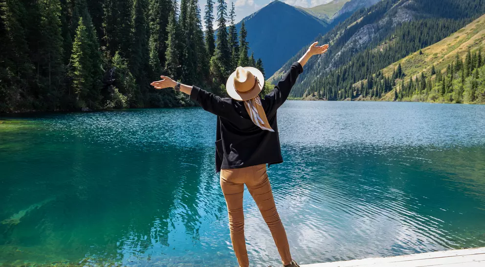 Person in a hat standing by a serene blue lake, arms outstretched, surrounded by trees and mountains under a clear sky.