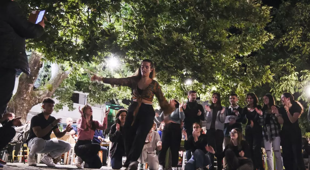 A lively outdoor scene with a dancer performing, surrounded by an engaged audience, under bright lights and trees.