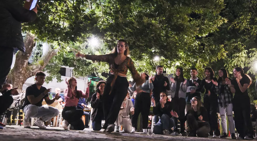 A lively outdoor scene with a dancer performing, surrounded by an engaged audience, under bright lights and trees.