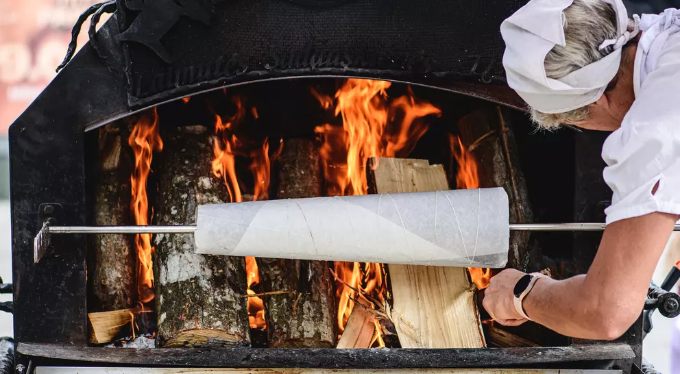 A person adjusts a cooking spit inside a wood-fired oven, with flames and logs visible in the background.