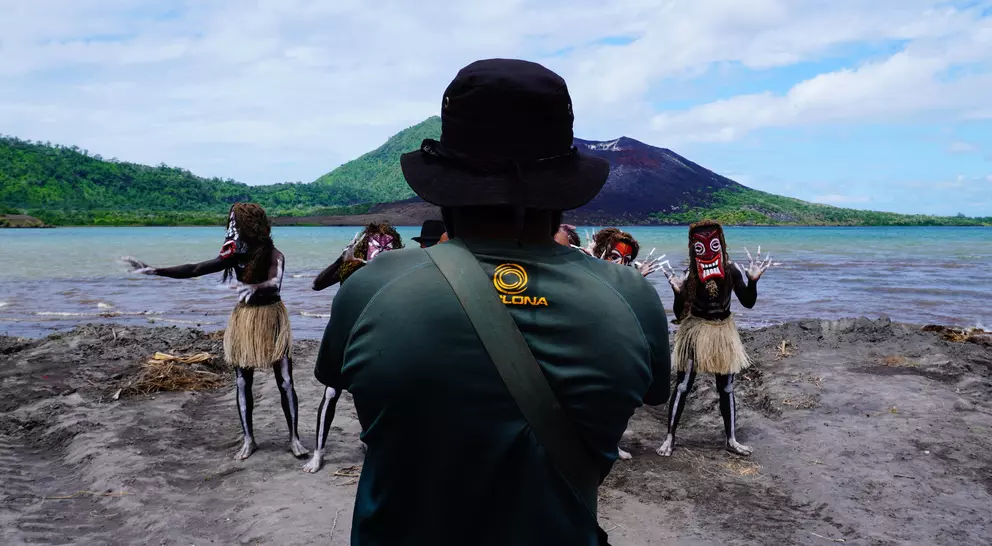 A person in a hat watches performers in masks and skirts dance by a serene waterfront and mountain backdrop.