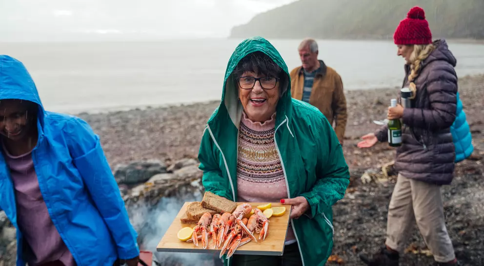 A smiling woman in a green jacket holds a tray of seafood by the beach, with friends nearby in casual attire.
