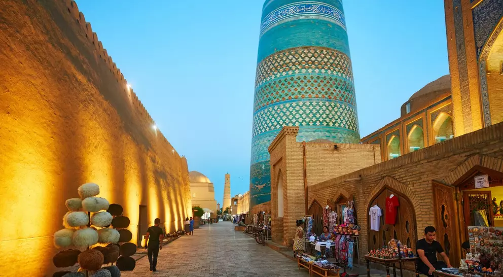 Evening scene in Khiva, Uzbekistan, showcasing a historic street lined with shops and a prominent blue minaret.