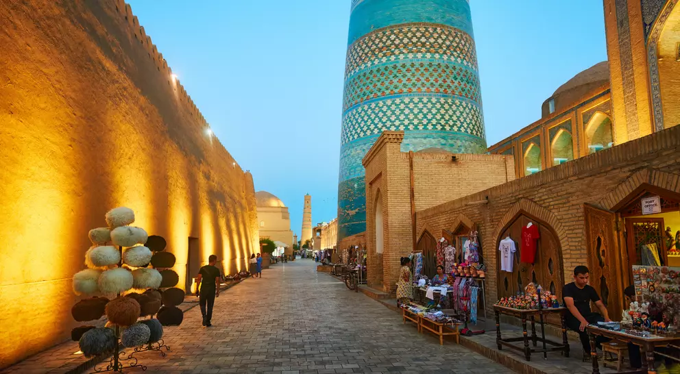 Evening scene in Khiva, Uzbekistan, showcasing a historic street lined with shops and a prominent blue minaret.