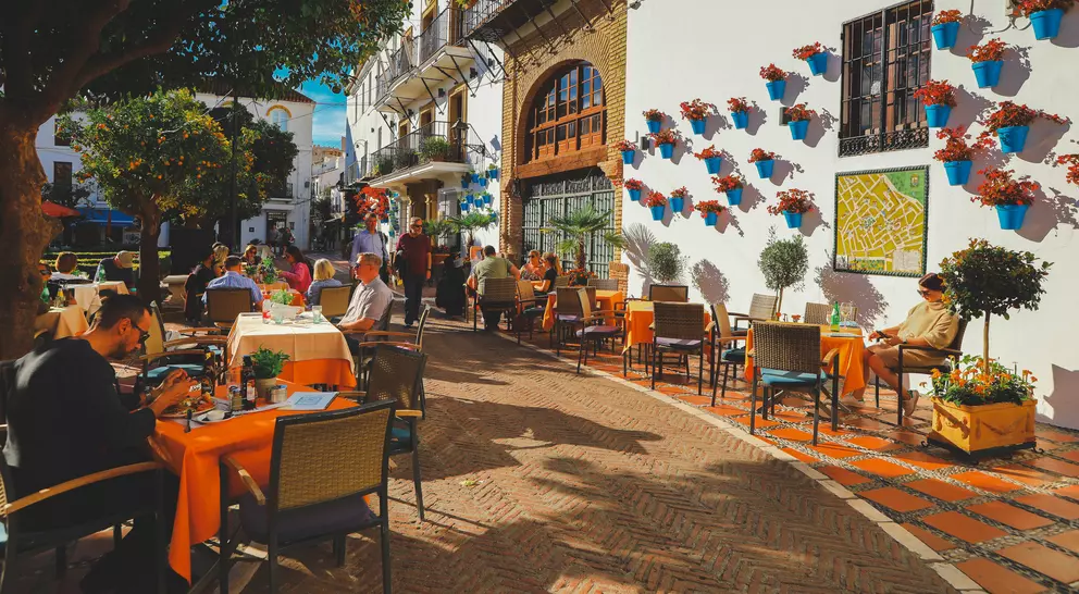 Outdoor café scene with people dining at tables, surrounded by vibrant flowers and a charming white building.