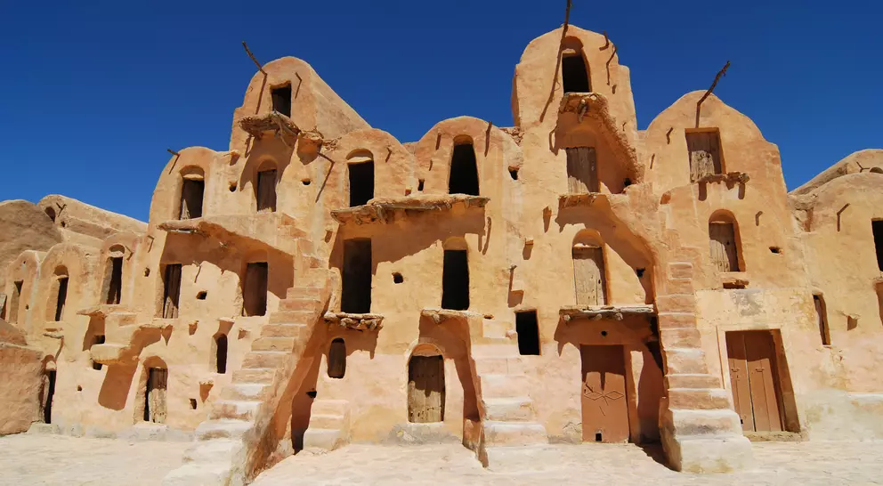 Ancient desert architecture featuring multi-level, ochre-colored buildings with stairs and numerous windows under a blue sky.