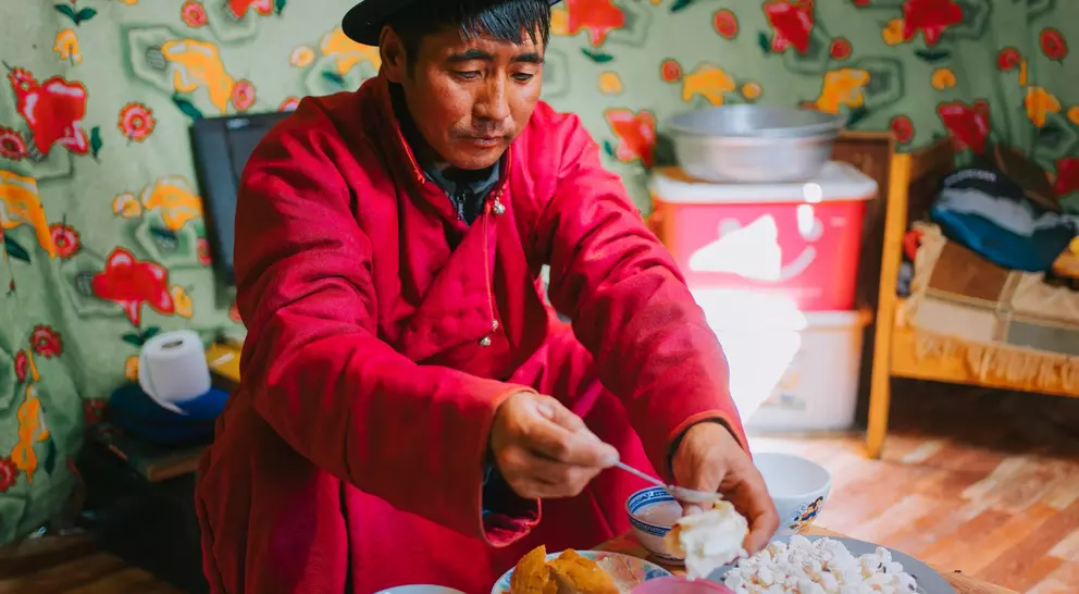 A man in a red robe serves food from a plate while sitting indoors, surrounded by colorful patterned walls.