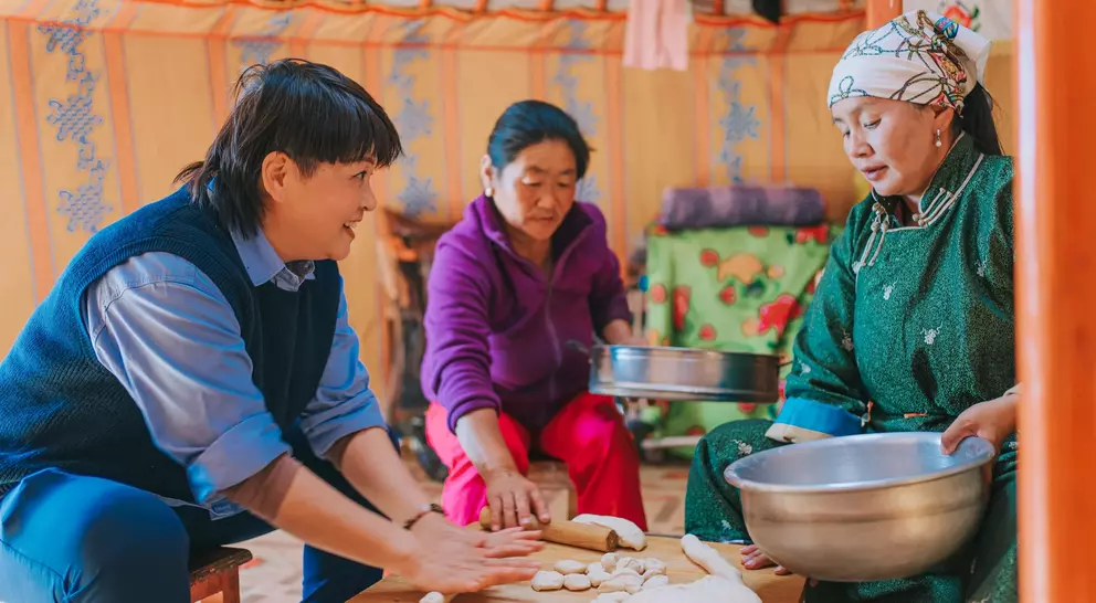 Three people prepare food inside a traditional yurt, with one person rolling dough and the others assisting at a table.