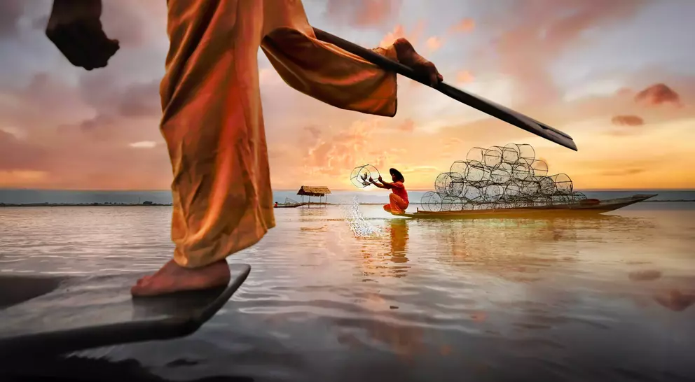 A fisherman in traditional dress uses a spear, while a woman in a boat casts nets at sunset over calm water.