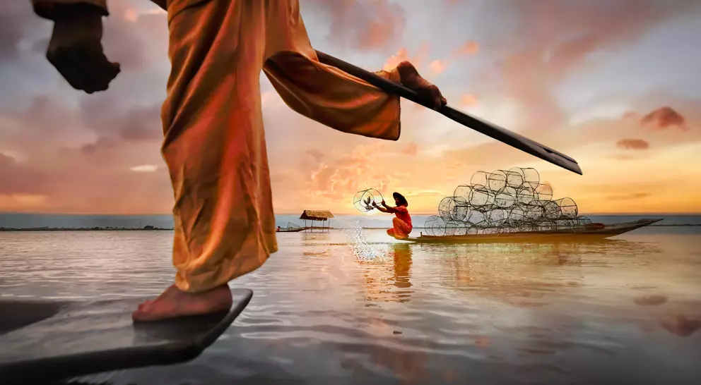 A fisherman in traditional dress uses a spear, while a woman in a boat casts nets at sunset over calm water.
