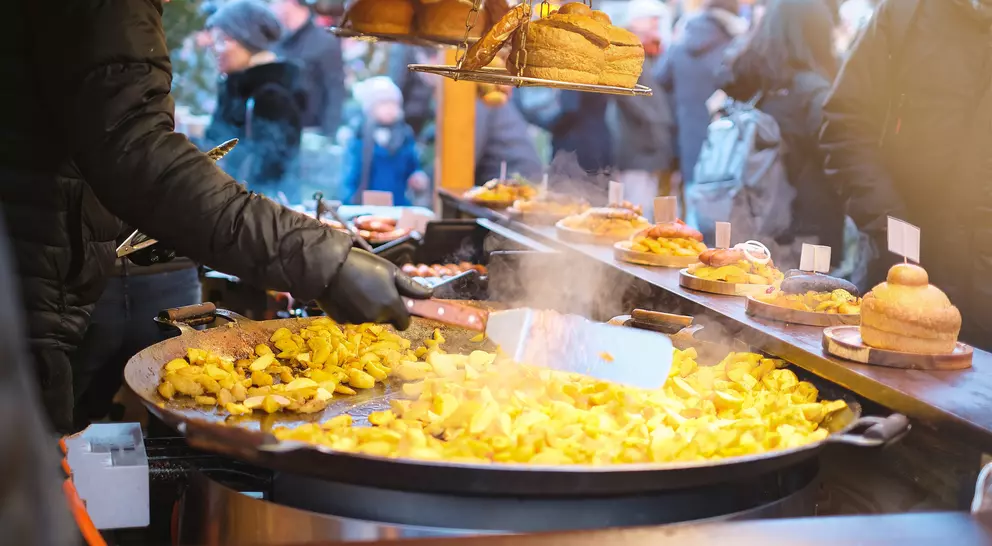 A vendor stirs a large pan of golden potatoes at a bustling outdoor market, surrounded by people and food stalls.