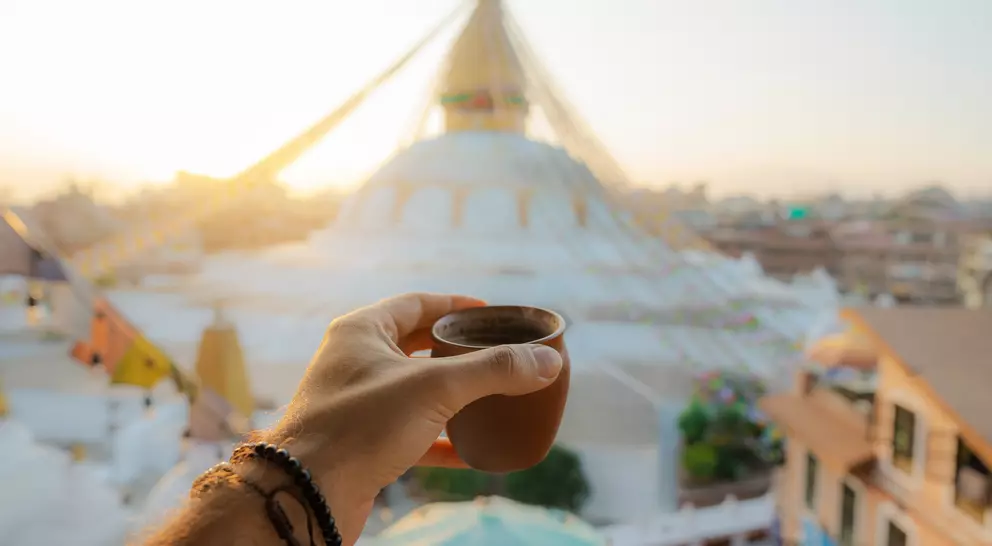A hand holding a small clay cup in front of a stupa at sunrise, with colorful prayer flags in the background.