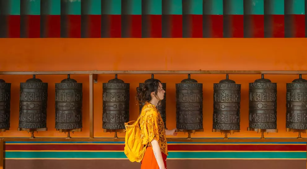 A woman in a yellow shirt and orange skirt walks past a row of prayer wheels against a vibrant orange and turquoise wall.